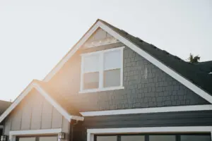 Sunlight shines on the upper portion of a modern house with dark grey siding, white trim, and a gabled roof. The image focuses on the attic windows and architectural details near the roofline.