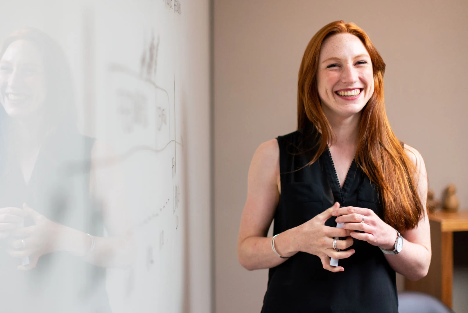 A woman with long red hair, wearing a sleeveless black top, stands smiling near a whiteboard with writing on it in an office setting. Her confident stance reflects the spirit of the Community Heroes program.