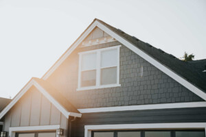 Sunlight shines on the upper portion of a modern house with dark grey siding, white trim, and a gabled roof. The image focuses on the attic windows and architectural details near the roofline.
