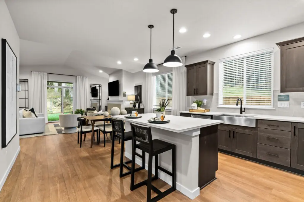 Modern open-concept kitchen and living area at Ponderosa Ridge with wood floors, dark cabinets, white countertops, a center island with stools, and a dining table; brightened by large windows and pendant lights.
