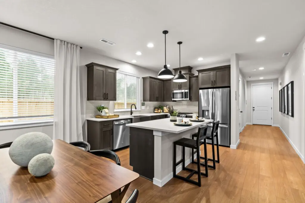 Modern kitchen in Ponderosa Ridge with dark cabinets, stainless steel appliances, a white island with three black stools, and pendant lights. Adjacent dining area features a wooden table and large windows with white blinds and curtains.
