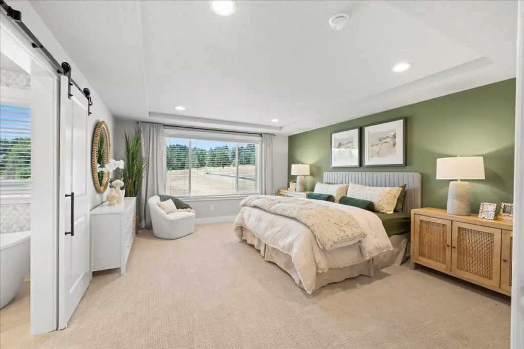 A cozy bedroom in the Pleasant Valley Villages housing development features a large bed with white and beige bedding. The green-painted walls complement the wooden bedside tables and lamps. The window offers a view of trees, while a white cabinet stands near the sliding door, adding charm to this real estate gem.