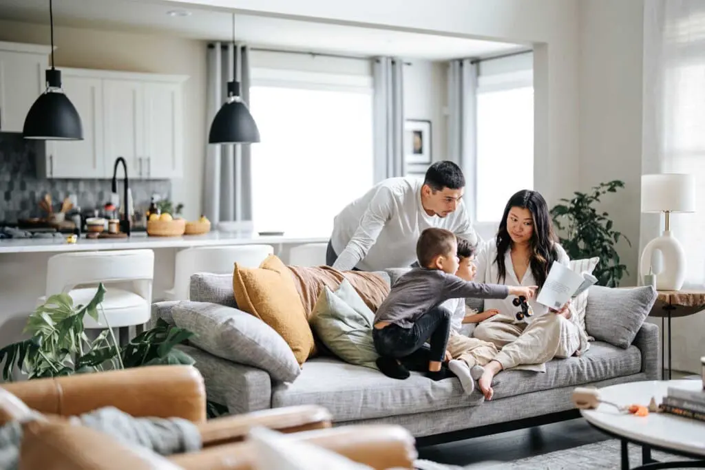 A family of four sits on a comfortable gray sofa in a modern living room. The mother is reading a book to two young children, while the father looks on. The room is bright with natural light, featuring a kitchen in the background.