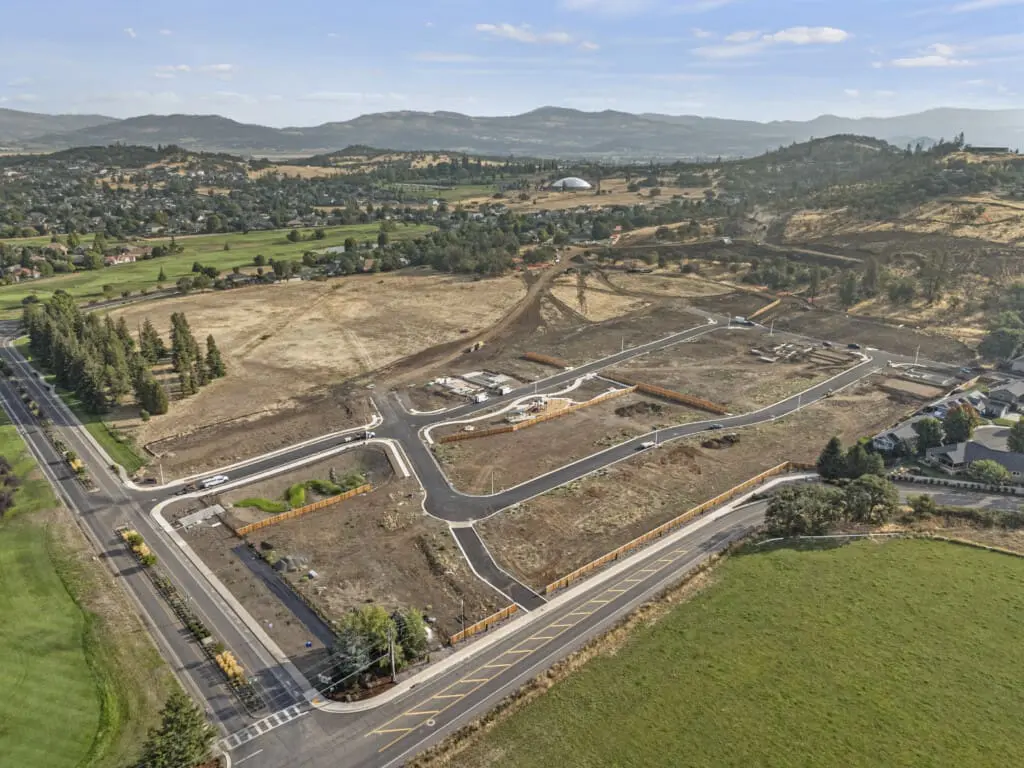 Aerial view of a rural area under development, featuring newly paved roads and cleared land. Surrounding the site are green fields, trees, and houses. Rolling hills and mountains are visible in the background under a clear blue sky.
