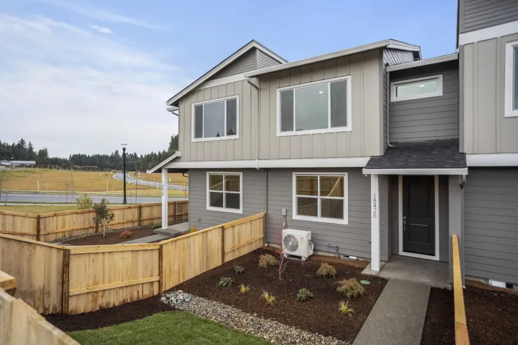 Located on NE 46th Place, this modern, two-story townhouse with gray siding and large windows features a small, landscaped front yard adorned with plants and a wooden fence. A path winds to the black front door. The surrounding area invites a tranquil ramble along roads and grassy fields under a cloudy sky.