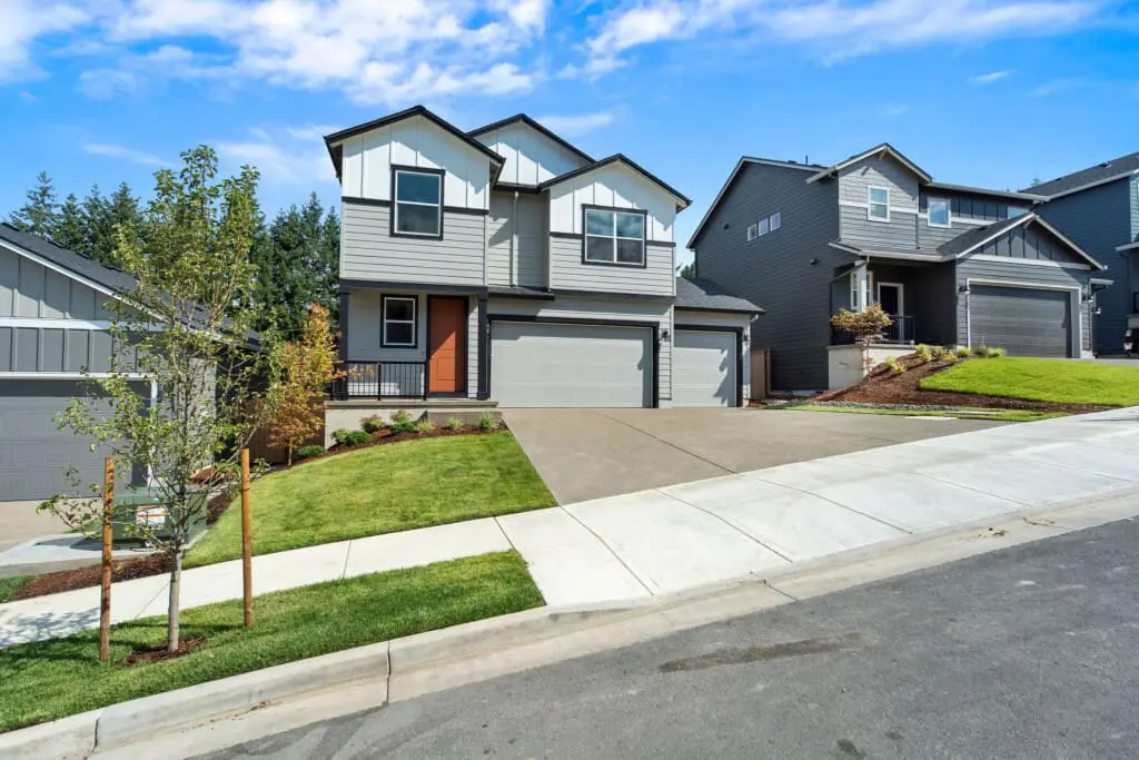 A modern two-story gray house with white trim and a red front door sits along NE Josephine Court in Faraday Hills, featuring a sloped driveway, green lawn, neighboring homes, and a clear blue sky.
