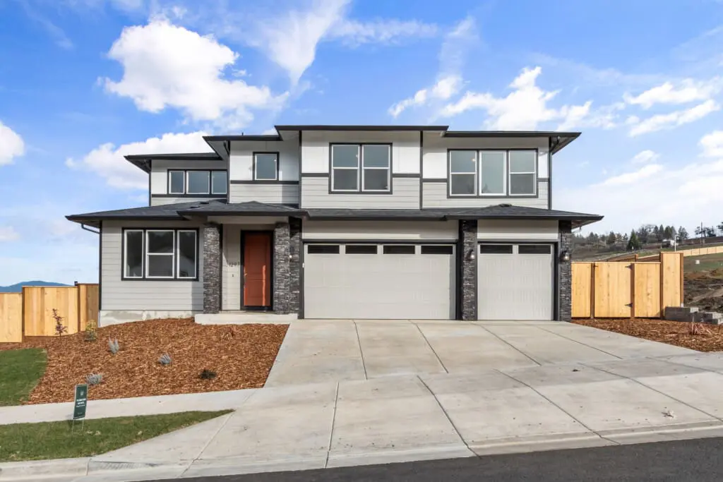 Modern two-story house with gray siding, large windows, and a three-car garage on Stonegate Drive, set on a newly landscaped yard with a wooden fence and concrete driveway under a partly cloudy sky.