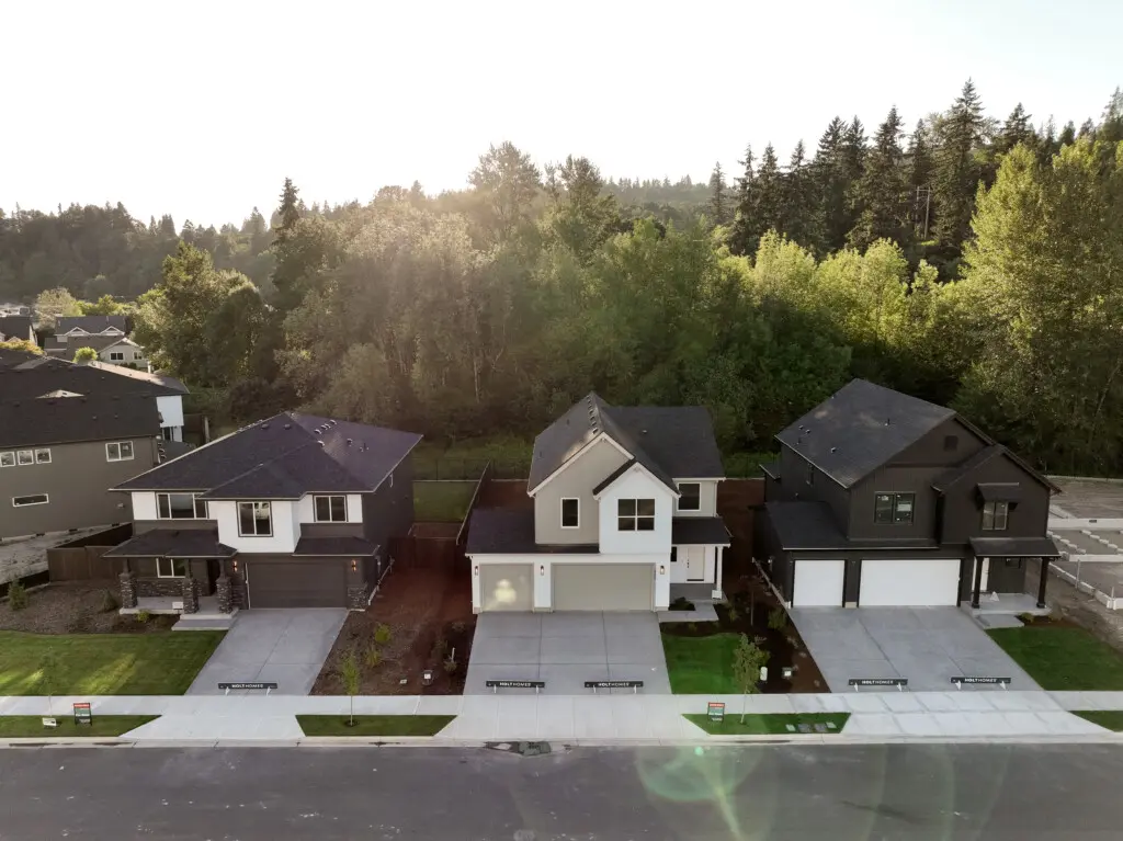 Aerial view of three modern, two-story suburban houses with driveways, garages, and lawns—showcasing right sizing and new home plans—set against dense green trees and rolling hills beneath a bright sky.