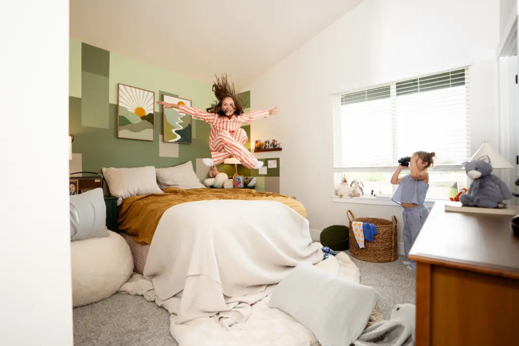 A child in striped pajamas jumps energetically off a bed in a cheerful, sunlit bedroom, while another stands near the window holding a stuffed animal. Art, toys, and cozy bedding fill the room with PNW charm reminiscent of local hiking trails.