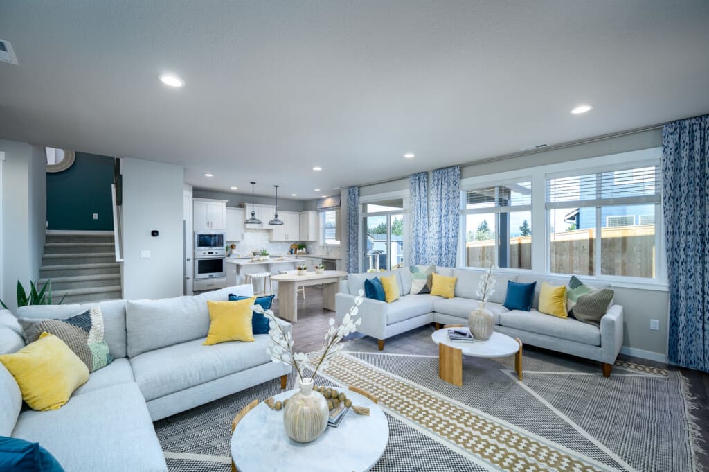 A modern, open-plan living room with two gray sofas, yellow and blue cushions, a patterned rug, and a round marble coffee table overlooking the serene East Mountain. The space flows into a kitchen with white cabinetry and a central island. Large windows allow natural light to fill the room.