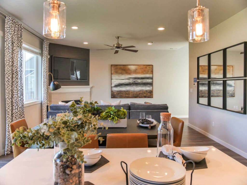 A modern living room and dining area with a gray couch, wall-mounted TV, abstract painting, brown accents, and large windows. The dining table has plants and decorative items, while pendant lights hang from the ceiling.