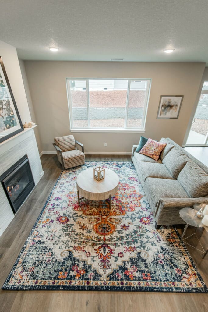 A living room with a large, patterned area rug, a gray sofa with colorful pillows, and a round wooden coffee table. Theres a fireplace on the left and a single armchair. Large windows allow natural light in, illuminating the space.