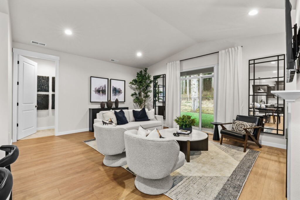 Modern living room at Ponderosa Ridge with light-colored walls, wooden floor, a cozy white sofa, two swivel chairs, a black accent chair, coffee table, large windows with curtains, wall art, and indoor plants. Natural light fills the space.