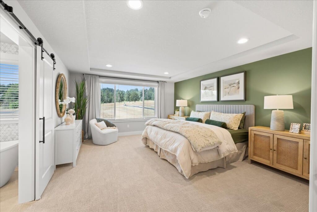 A cozy bedroom in the Pleasant Valley Villages housing development features a large bed with white and beige bedding. The green-painted walls complement the wooden bedside tables and lamps. The window offers a view of trees, while a white cabinet stands near the sliding door, adding charm to this real estate gem.