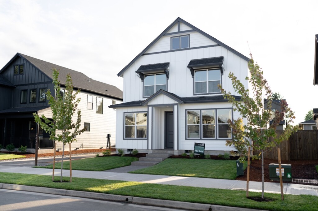 A modern two-story white house with large windows, a front porch, manicured lawn, young trees, and a For Sale sign—ideal for first time home buyers in Oregon—nestled in a welcoming suburban neighborhood.
