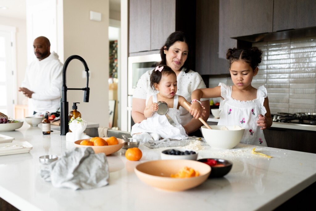 A woman and two young girls bake together in a modern kitchen, possibly creating memories as first time home buyers in Oregon. One girl stirs a bowl, the other holds a measuring cup; fruits and bowls fill the counter while a man stands in the background.