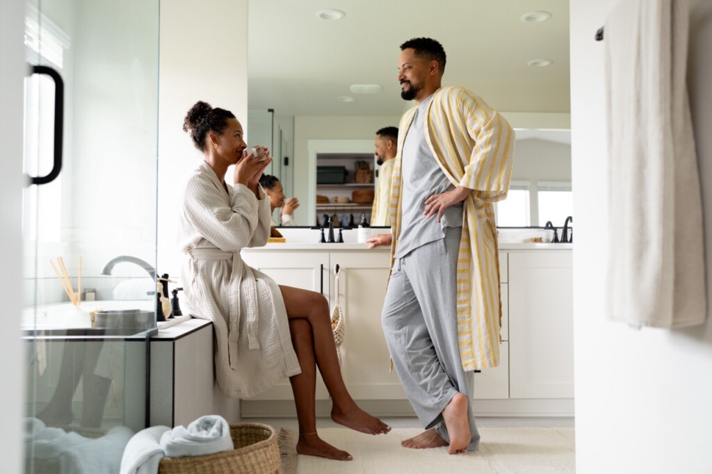 A woman sits on a bathroom counter drinking from a mug while a man in pajamas and a yellow-striped robe stands nearby; both first time home buyers in Oregon, smiling and talking in their bright, modern bathroom.