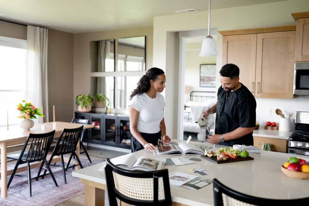 A smiling couple, first time home buyers in Oregon, stands in a bright kitchen. One flips through a book on the island counter while the other pours water, surrounded by fresh fruits, vegetables, and kitchenware. Plants and the dining table are visible behind them.