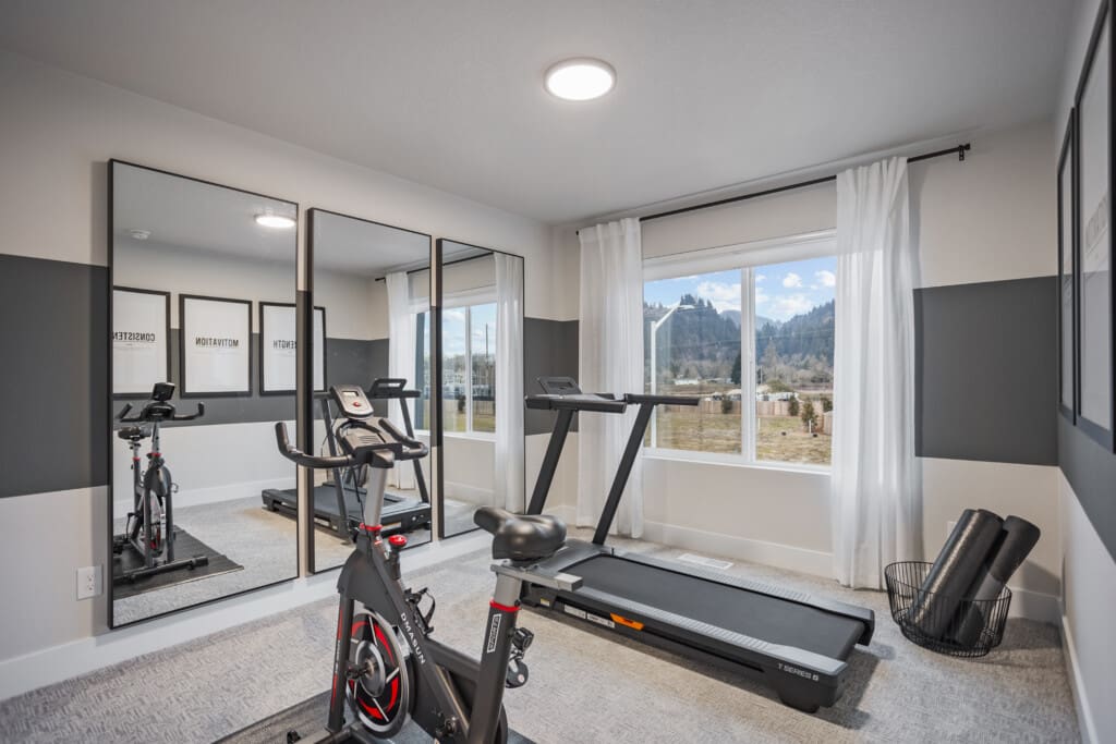 Home gym with a stationary bike and treadmill facing large mirrors. Framed motivational quotes line the wall. A window with white curtains reveals a scenic mountain view. Yoga mats are rolled in a basket in the corner.