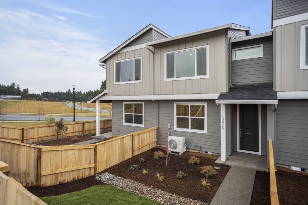 Located on NE 46th Place, this modern, two-story townhouse with gray siding and large windows features a small, landscaped front yard adorned with plants and a wooden fence. A path winds to the black front door. The surrounding area invites a tranquil ramble along roads and grassy fields under a cloudy sky.