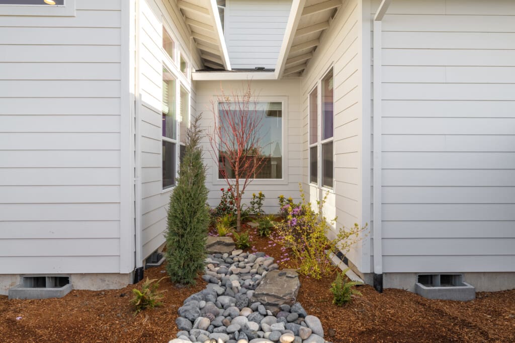 A modern house exterior with white paneling features a small landscaped garden. The garden includes a dry riverbed made of smooth stones and is surrounded by various small shrubs and plants. A window is centered in the background.