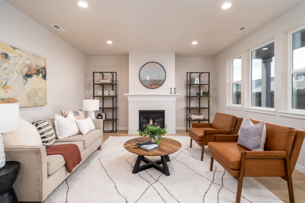 Modern living room at Greely Farms with a beige sofa, two brown chairs, and a round wooden coffee table on a geometric rug. A fireplace with a round mirror above is flanked by black shelves holding decorative items. Large windows at 8700 S 1st St let in natural light.