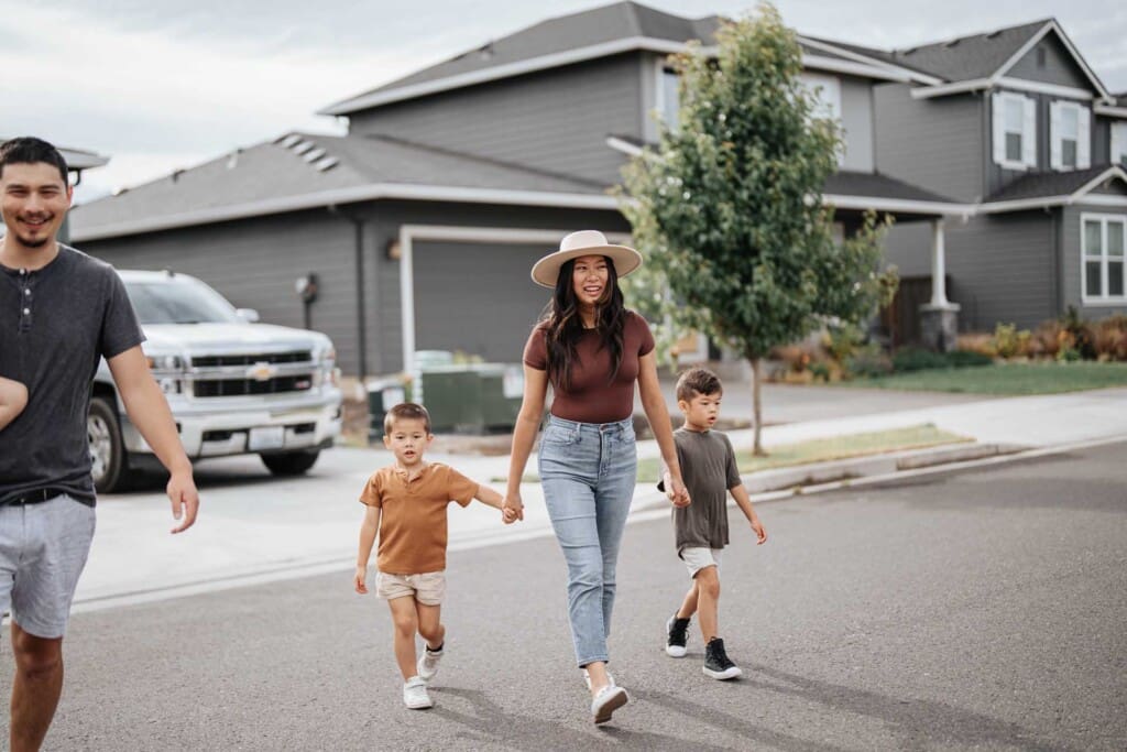 A woman holding hands with two young boys walks across a suburban street, while an adult man walks nearby. The group is in front of modern gray houses on a cloudy day.