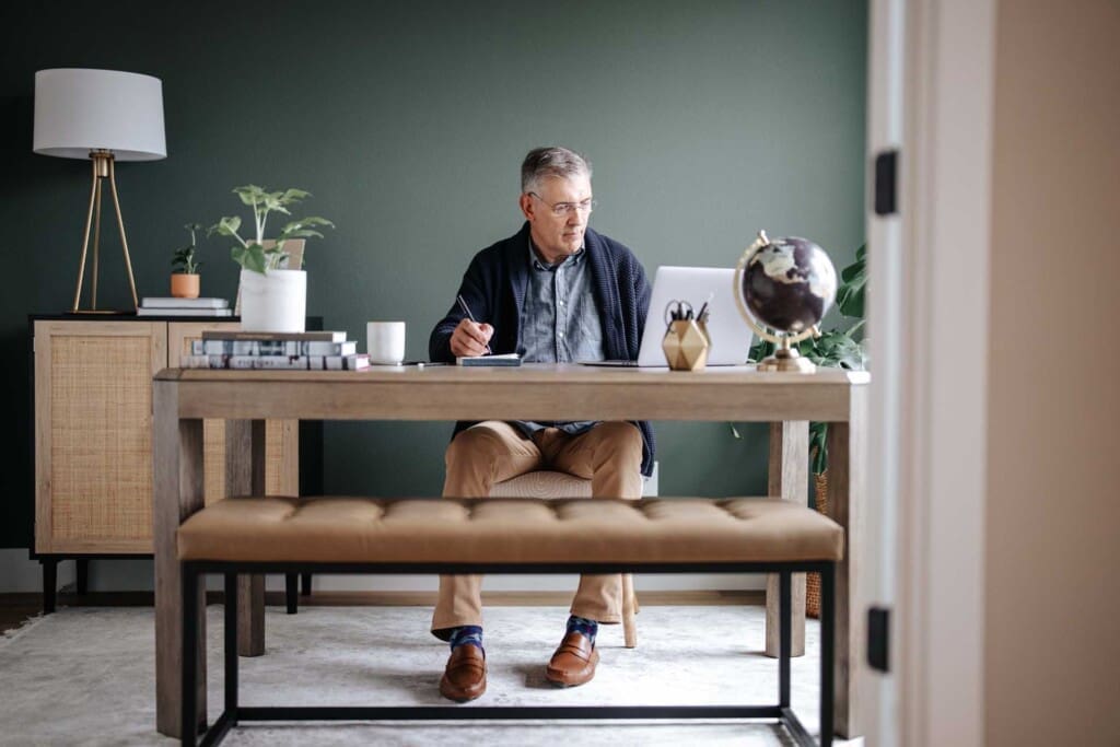 An older man sits at a desk in a home office, writing in a notebook while looking at a laptop. The desk has a globe, pencils, books, and a mug. The room is decorated with plants and modern furniture.
