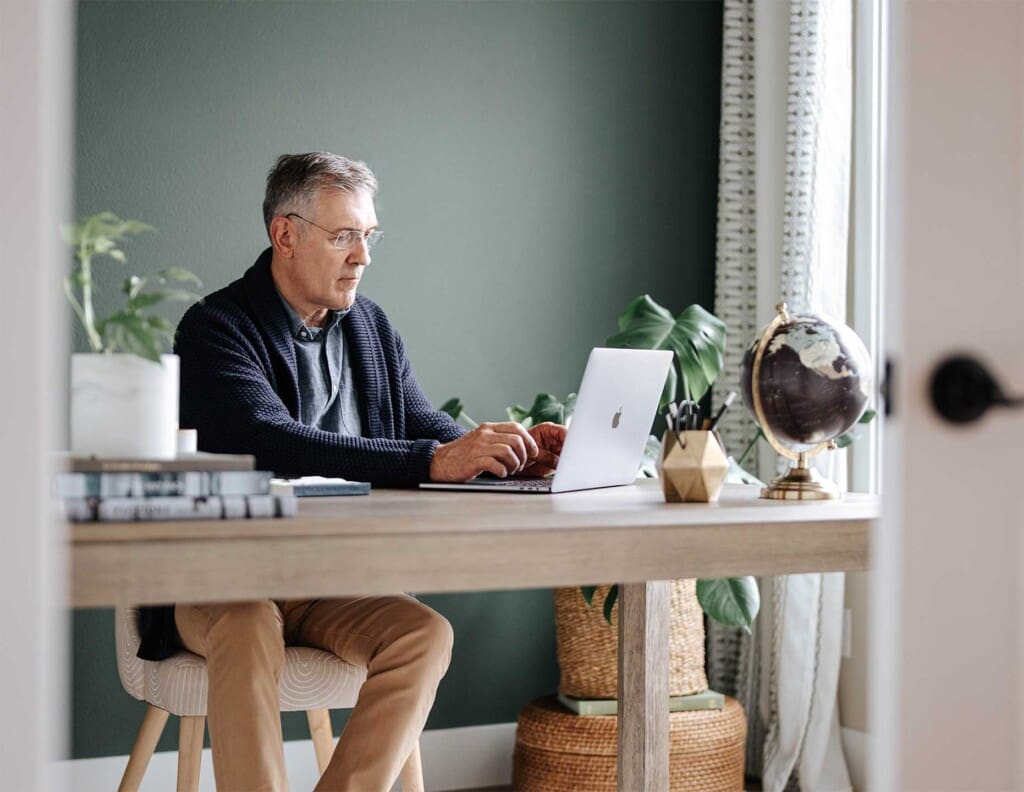 An older man sits at a modern desk working on a laptop in a bright, stylish home office with plants, books, and a globe.