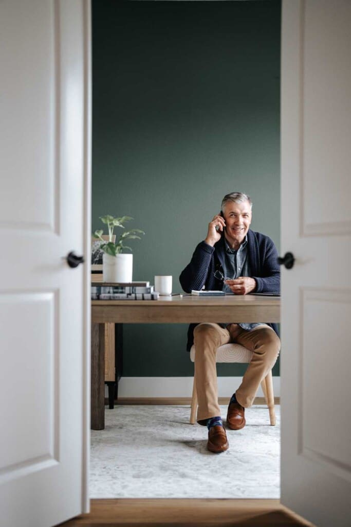 A man sits at a desk in a home office, talking on a smartphone. The room has a green wall, a plant, a mug, and books on the desk. He appears relaxed and is smiling. The scene is viewed through open double doors.