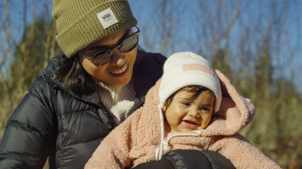 A woman wearing sunglasses and a green beanie smiles while holding a baby dressed in a pink fuzzy jacket and white beanie. They enjoy a sunny day together at Willow Ridge, surrounded by trees in the background.