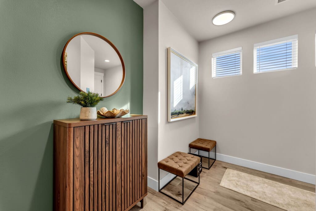 A modern Harmony Heights entryway with a wooden cabinet, round mirror above it, small plant decor, two brown tufted benches, a landscape painting, green accent wall, three small windows, and light wood flooring.