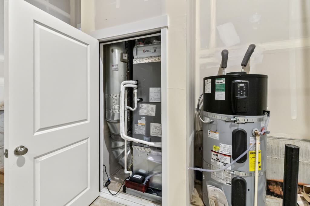 An open closet reveals an HVAC unit with exposed pipes and labels. Next to it stands a water heater with instructional stickers and pipes attached, all inside a partially finished utility room.