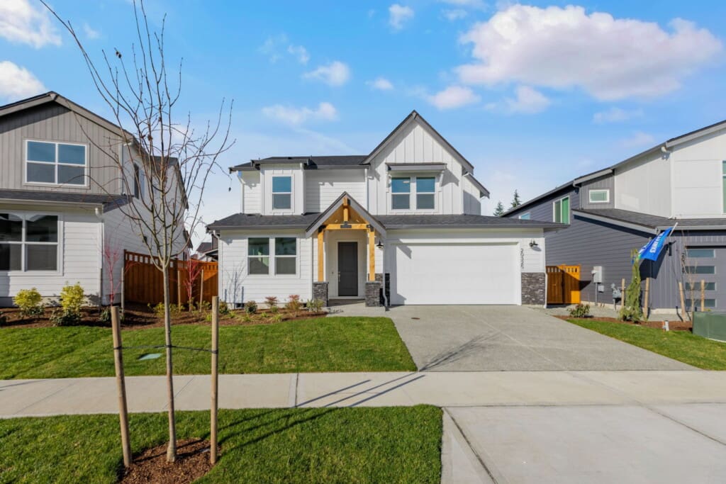 A modern two-story white house with a gray roof, stone accents, and a covered porch, set on a manicured lawn with a driveway, young tree, and neighboring homes under a bright partly cloudy sky.