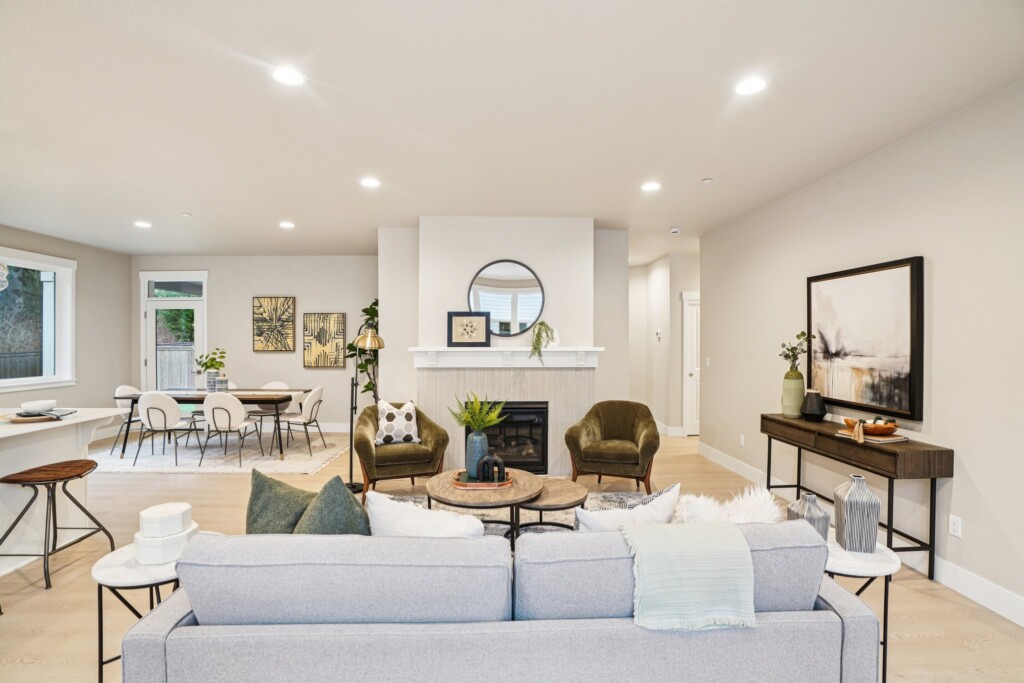 Modern living room with light walls, a gray sofa, two green chairs, a fireplace with a round mirror above it, and a console table with a large framed artwork. Dining area and kitchen visible in the background.