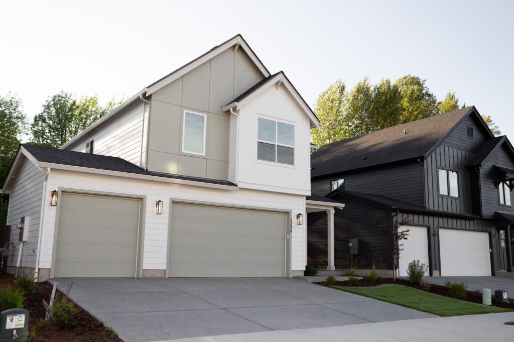 A modern two-story new construction home with a gray and white exterior, three-car garage, and neatly landscaped front yard sits next to a similarly styled dark gray house on a sunny day—perfect for anyone seeking a new home buying guide.