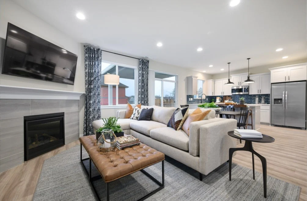 Tour this modern open-concept living room and kitchen, featuring the 1857 Model hearth, a beige sofa, brown tufted bench, wall-mounted TV, fireplace, dining table, large windows, wood floors, and stylish decor.