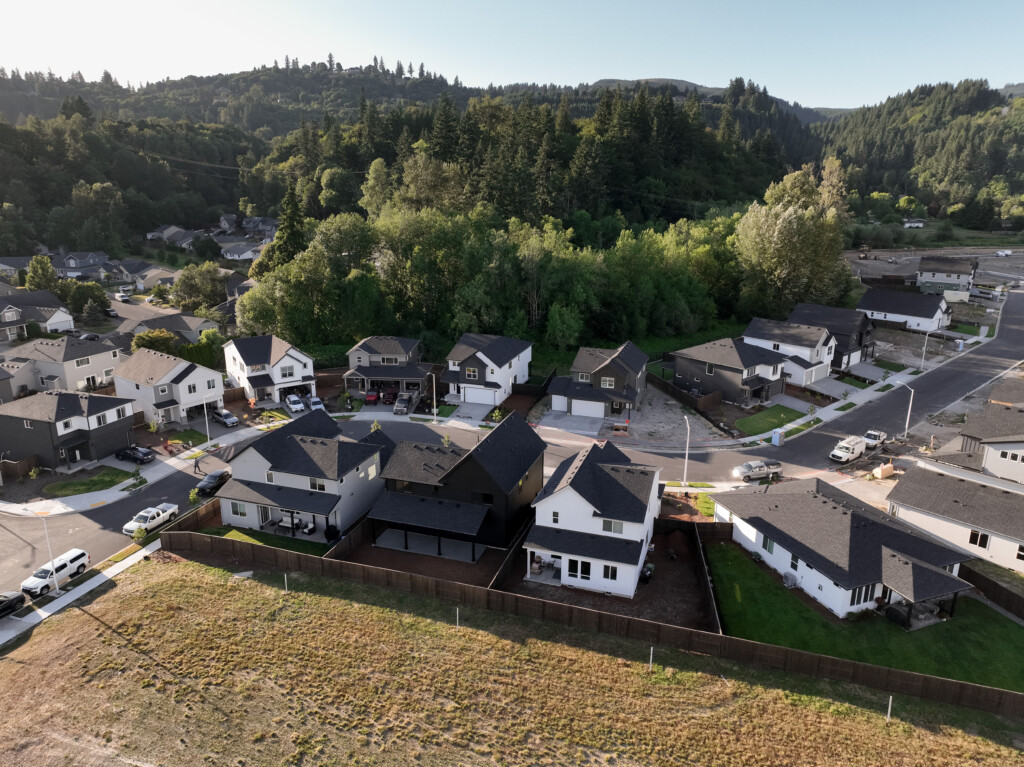 Aerial view of a suburban Portland, Oregon neighborhood with modern houses, green trees, and hills in the background on a clear day. The calm area boasts new homes, neat yards, and open land—perfect for exploring local things to do.