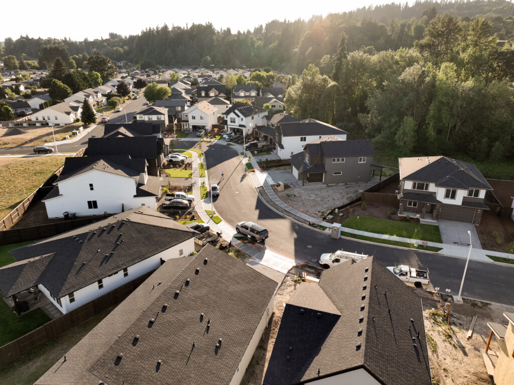 Aerial view of a modern suburban neighborhood in Woodland WA with rows of houses, tree-lined streets, parked cars, and a wooded area in the background under a bright sky.