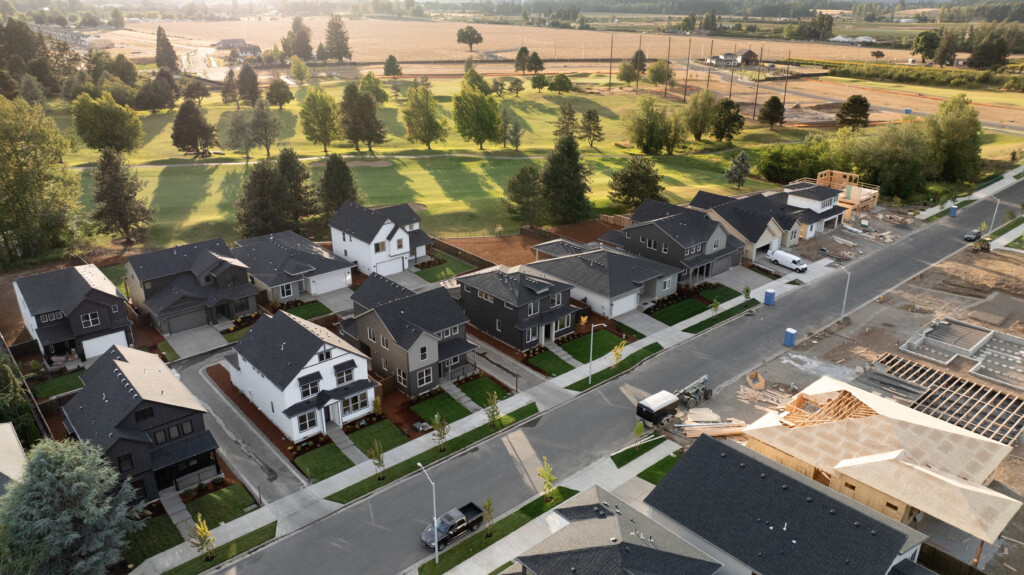 Aerial view of a suburban neighborhood in Tehaleh, Washington, with modern houses, manicured lawns, and parked cars. A green park with trees sits behind the homes, typical of the lush PNW landscape; new construction appears on the right side.