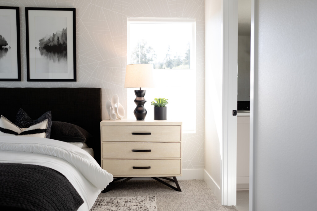 A modern bedroom at The Hearth features a neatly made bed with black and white bedding, a nightstand with three drawers, a lamp, a plant, and decorative objects. Sunlight streams through a window above the nightstand in the 1594 Model Home.