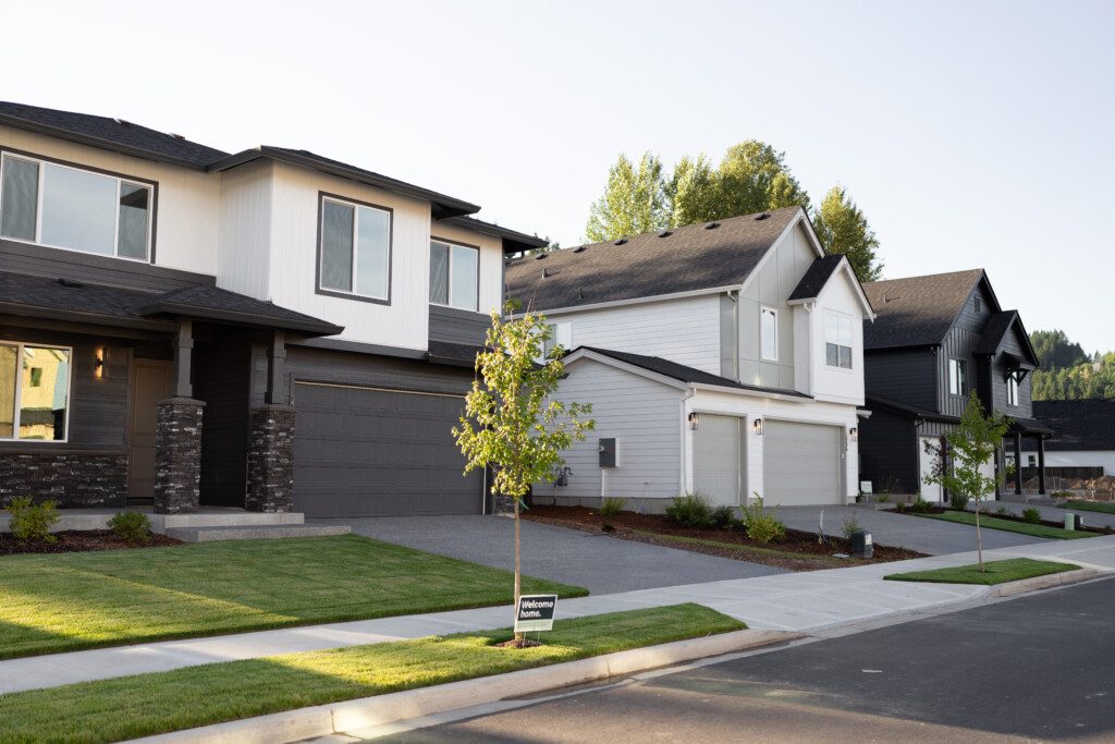 A row of modern two-story suburban houses with well-kept lawns, young trees, and driveway entrances, taken on a sunny day. A for sale sign is visible on one lawn, highlighting the home's 10-year structural warranty.