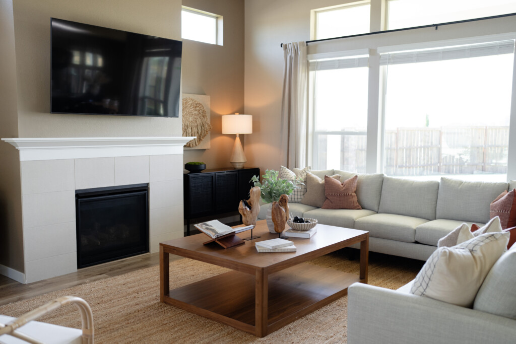 Modern living room with a wall-mounted TV above a fireplace, a light gray sectional sofa, wooden coffee table, lamp, and large windows—perfect for relaxing after exploring Southwest Washington or planning your next travel adventure.