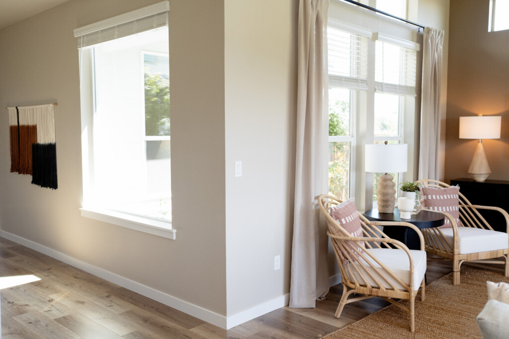 Bright living space with wooden chairs, pillows, a round table, and a lamp by large windows with sheer curtains. Ideal for home office makeovers, its airy atmosphere and cozy accents offer comfort during the colder months. Wall art hangs near another window.