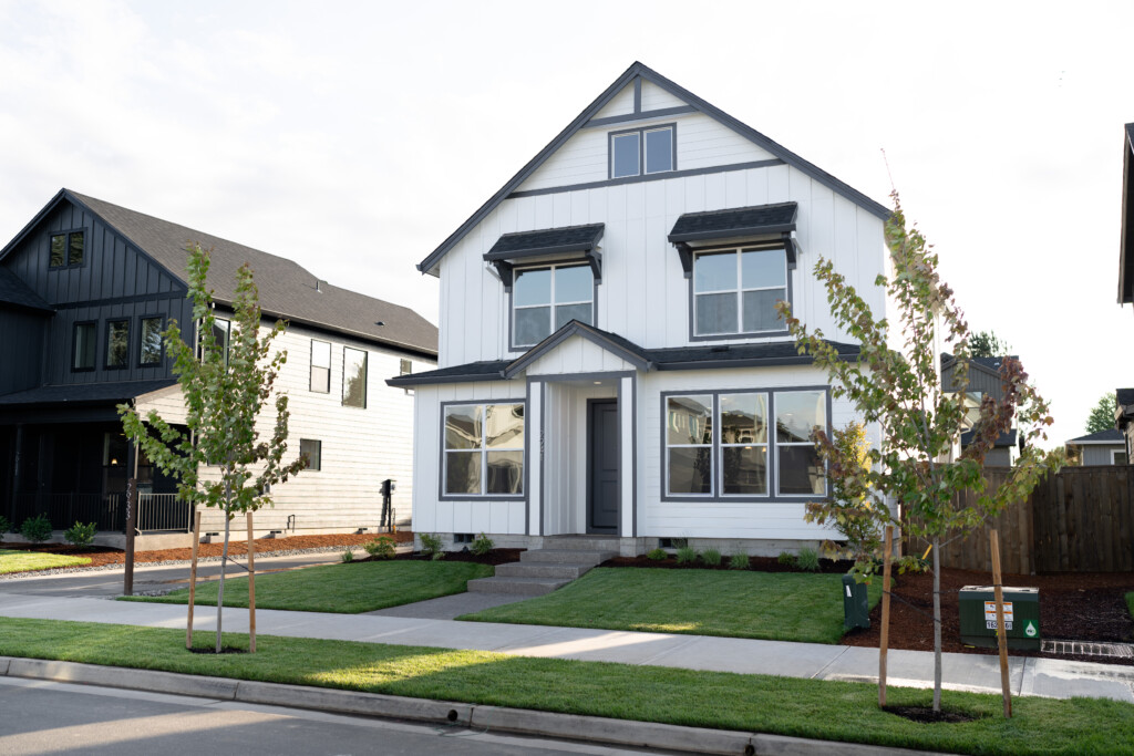 A modern two-story white house with black trim, large windows, and a front porch. Newly planted trees and freshly mowed grass line the sidewalk. Enjoy peace of mind with workmanship warranty coverage as neighboring homes are visible on either side.