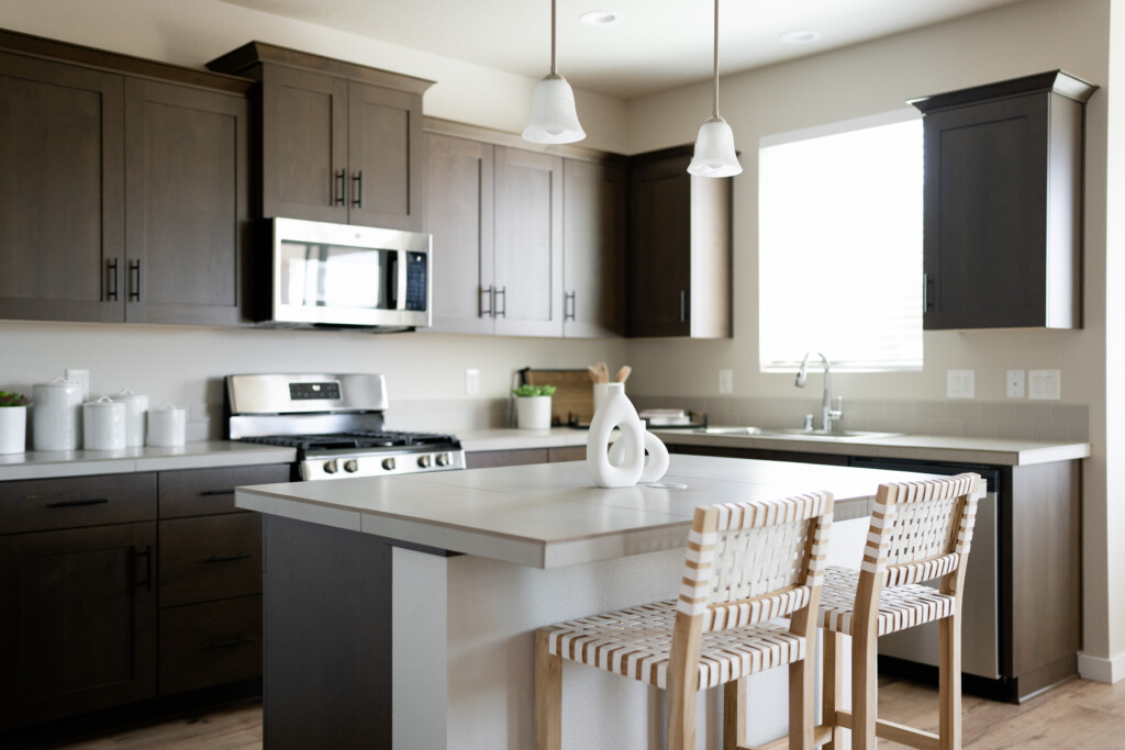 A modern new-build kitchen with dark cabinets, a stainless steel stove, built-in microwave, island with white countertop, two woven barstools, pendant lights, a window above the sink, and decorative accessories on the counters.