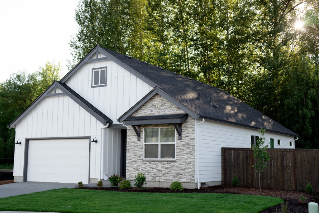 A modern single-story new construction house with white siding, stone accents, a gable roof, two-car garage, and a well-kept lawn, surrounded by a wooden fence and tall trees in the background.
