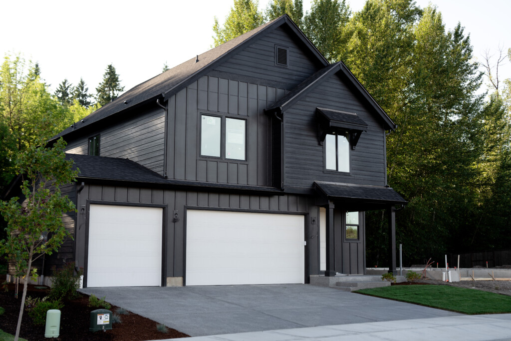 Modern two-story black house with white garage doors, a sloped roof, and large windows, surrounded by trees and expertly landscaped yard that’s easy to care for, plus a concrete driveway.