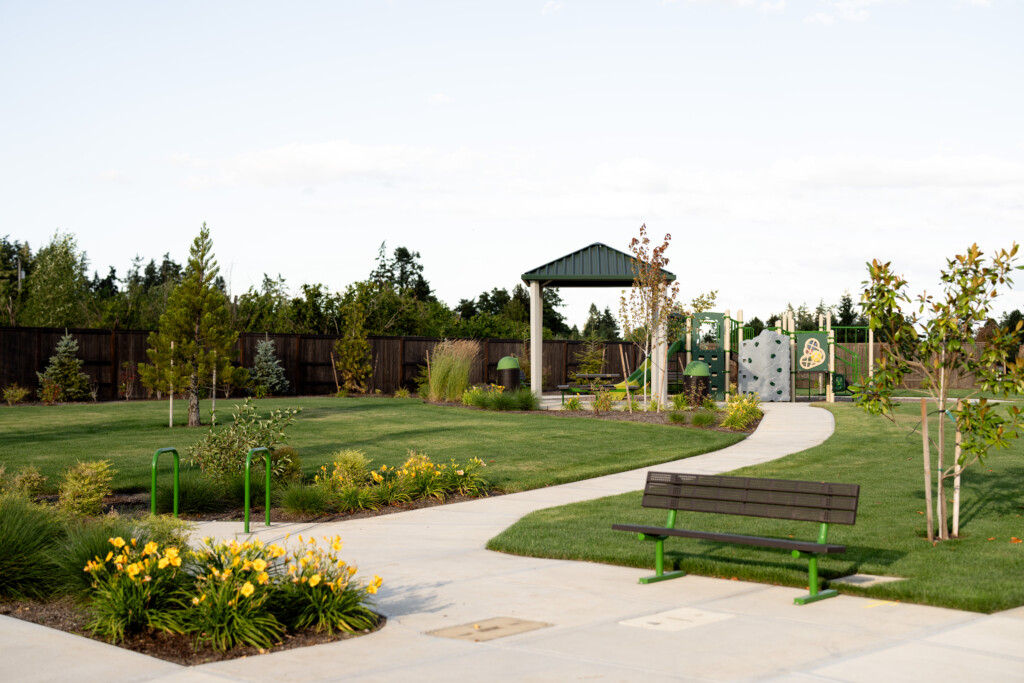 A park in Bonney Lake, Washington offers plenty of things to do, with a green lawn, yellow flowers, a paved walkway, a wooden bench with green legs, exercise bars, and a playground featuring a climbing wall and shaded pavilion.