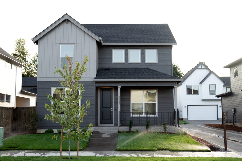 A modern two-story gray house with white trim, a dark front door, and a small tree in the front yard features large windows and a well-planned drainage system. It’s nestled in a suburban neighborhood with similar homes nearby.